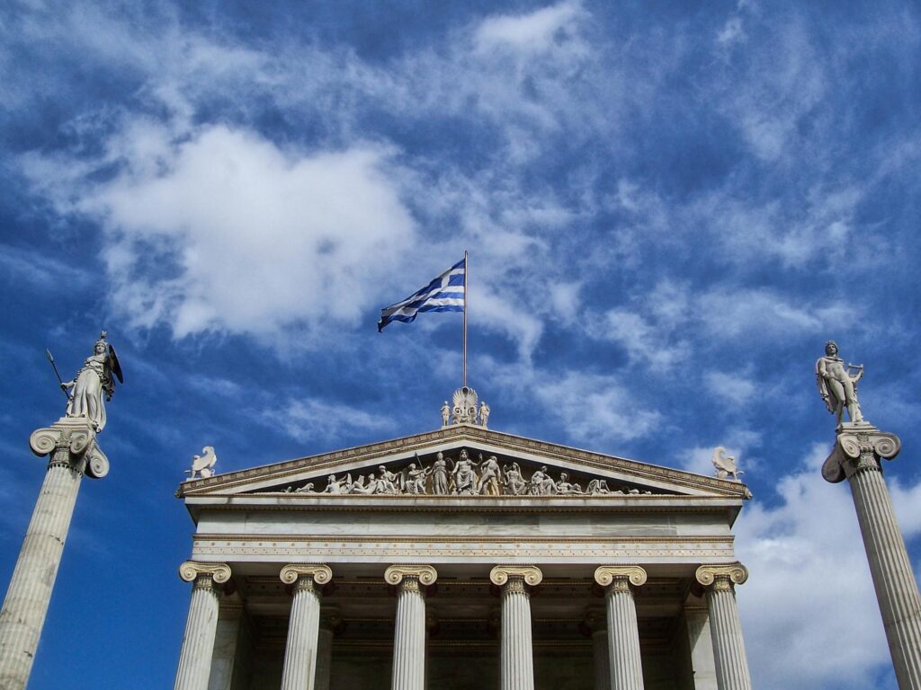 Front view of the Academy of Athens with Greek flag and statues against a vibrant sky.