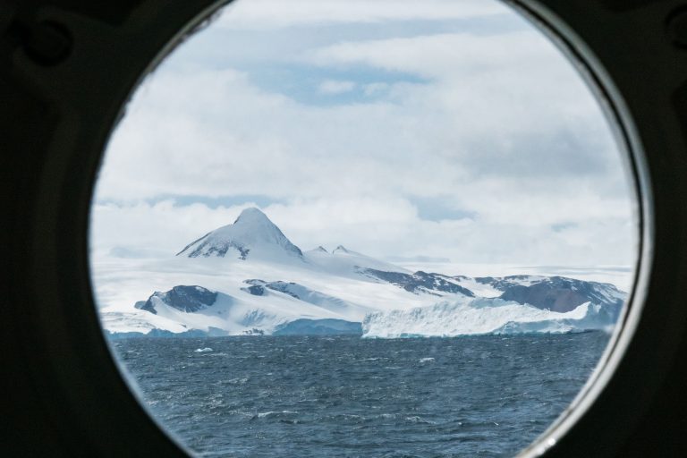 A stunning view of Antarctica's icy landscape through a ship's porthole, featuring snow-capped mountains and the open sea.