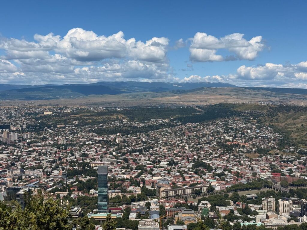 Aerial view of Tbilisi, Georgia, showcasing cityscape against mountain backdrop under a blue sky.